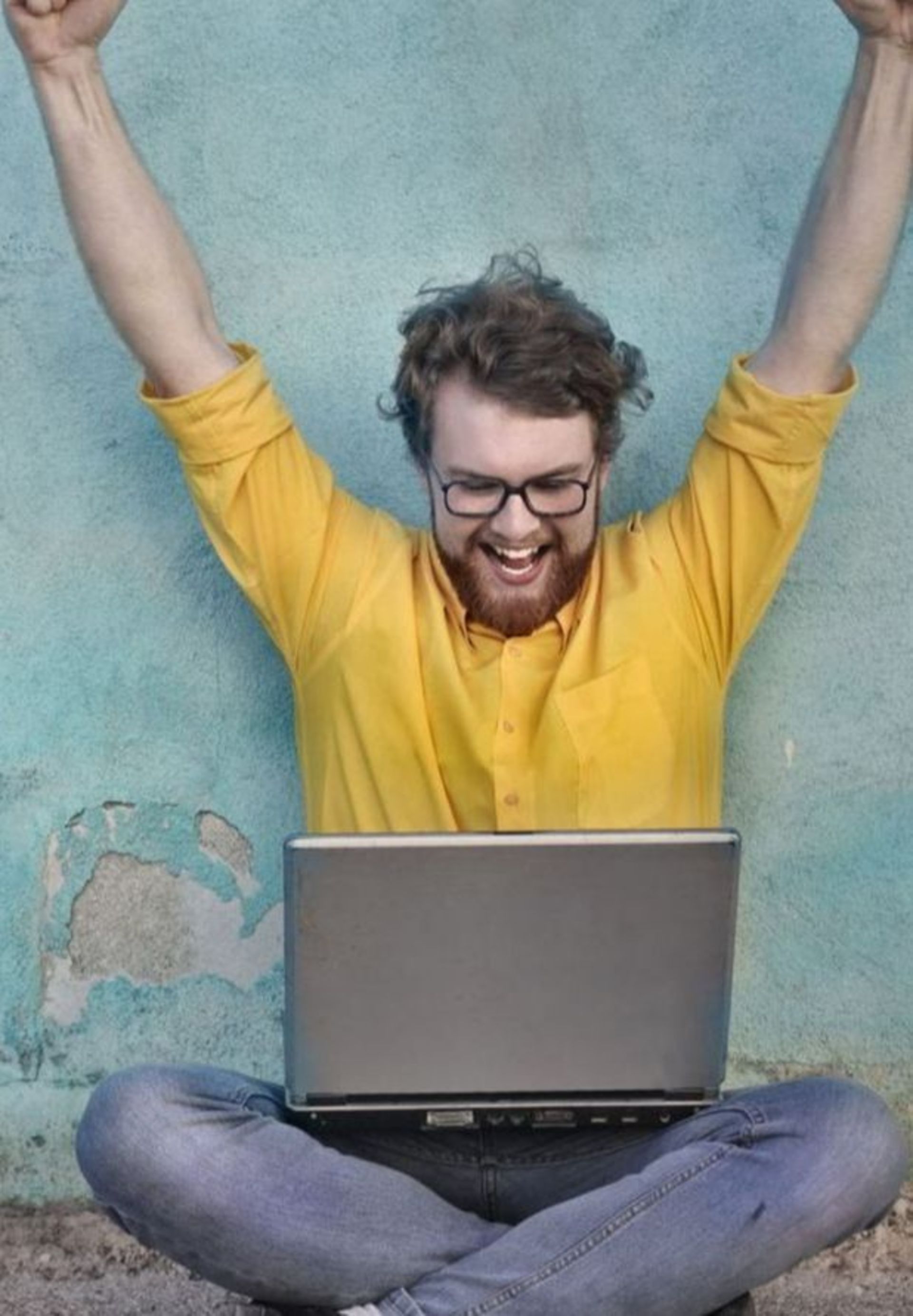 man sitting cross legged with laptop and arms triumphantly in the air 